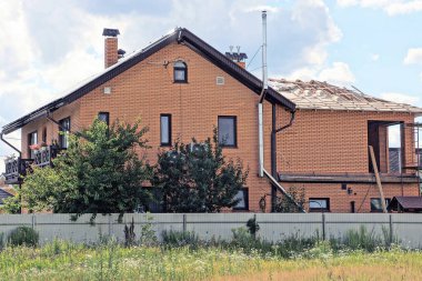 Gri metal bir çitin arkasında çatısı olan büyük kahverengi tuğladan bir ev. Gökyüzüne karşı yeşil çimenler ve bitki örtüsü.