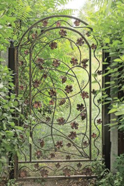 Metal çubuklardan yapılmış kapalı kahverengi bir kapı. Sokakta yeşil bitki örtüsünün arasında dövülmüş bir desen var.