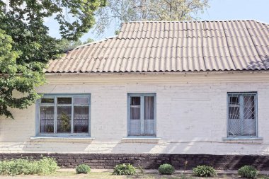 Kırsal bir sokakta yeşil bitki örtüsü arasında gri bir çatı altında pencereleri olan beyaz, eski bir tuğla evin ön cephesi.