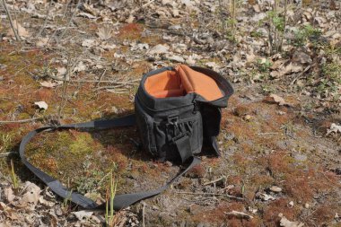 Doğada kahverengi yosun ve gri zemin üzerinde askısı olan açık kırmızı bir kumaş çantası var.