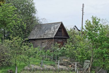 Gri bir çatı altında, yeşil bitki örtüsü ve ağaçların arasında, eski, kahverengi, ahşap bir kır evi gökyüzüne karşı.