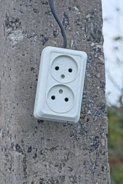 one plastic power strip with two electrical outlets with black wire hangs on a gray concrete pole in the street