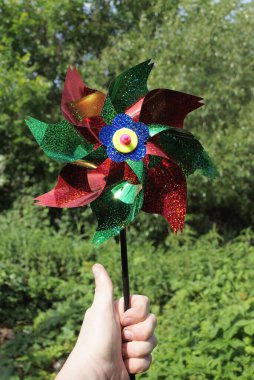 Doğadaki yeşil bitki örtüsünün arka planında büyük renkli plastik bir oyuncak yel değirmeni tutuyor.