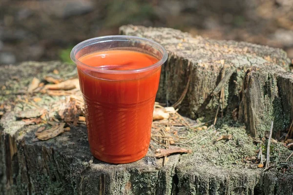 天然の灰色の切り株に赤いトマトジュースが入ったプラスチック製のガラスが立っています — ストック写真