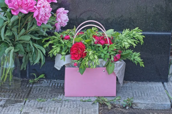 a bouquet of red rose flowers with green leaves in a pink bag stands on a gray sidewalk against a black marble wall in the street