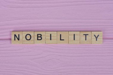 Küçük gri ahşap harflerden gelen soyluluk kelimesi pembe bir arka planda yatıyor.