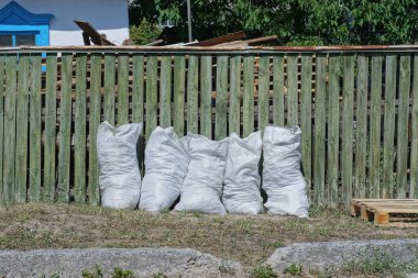 büyük beyaz bir sıra çöp dolu beş plastik torba yeşil ahşap bir çit duvarın yanında kırsal bir sokakta çimlerin üzerinde