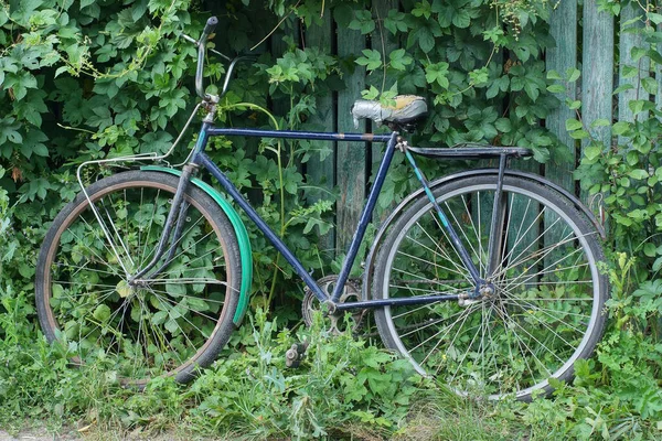 一辆蓝色的旧自行车靠在乡村街道上长满青草的木篱笆墙上 — 图库照片