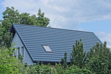 Gri kiremitli çatının altında, yeşil ağaçların arasında tek penceresi olan ve gökyüzü ve bulutların arka planına karşı sokakta bulunan özel bir evin tavan arası.