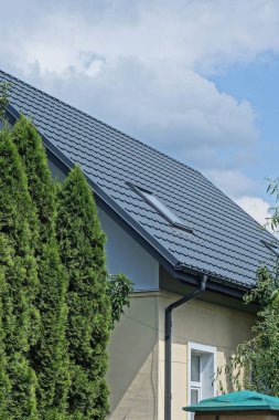 Gri kiremitli çatının altında, yeşil ağaçların arasında tek penceresi olan ve gökyüzü ve bulutların arka planına karşı sokakta bulunan özel bir evin tavan arası.