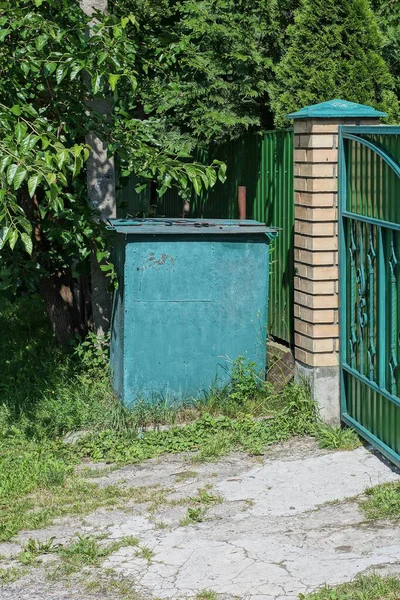 one green closed metal trash can stands against the wall of the fence in the grass on the street
