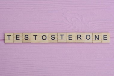 Küçük gri ahşap harflerden gelen testosteron kelimesi pembe ahşap bir masada yatar.