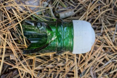 Kırık bir yeşil cam şişe boynu, beyaz plastik bir kapakla sokaktaki gri kuru çimenlerin üzerinde yatıyor.