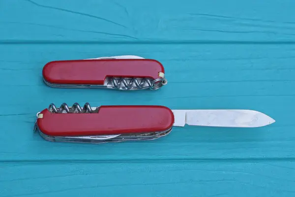two open folding knives multitools with red handles with gray blades lie on a blue green wooden table