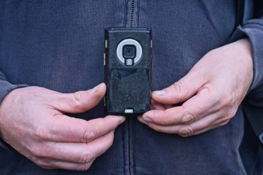 Gri elbiseli bir adamın ellerinde eski siyah bir cep telefonu ve caddede küçük bir kamera lensi var.