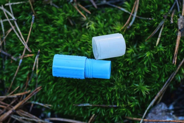 one small open blue plastic tube and a white cap lie on green moss on the street