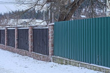 Kahverengi ahşap tahtalardan, taşlardan ve beyaz karda yeşil metalden yapılmış bir çit parçası.