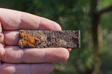 Kahverengi, küçük, paslı bir levha yeşil arka planda bir elin avucunda yatar.