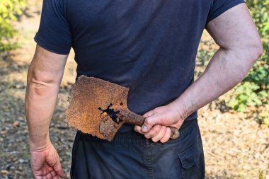Siyah elbiseli bir adam, bir yaz caddesinde elinde paslı, eski bir demir siper aletini tutuyor.