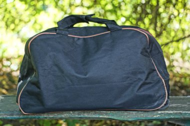 Bir yaz caddesinde bir bankta yeşil bitki örtüsünün arka planında duran büyük siyah bir çanta.