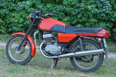 Eski bir kırmızı siyah motosiklet yerde ve yeşil çimenler yaz caddesinde duruyor.