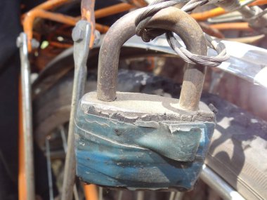 Bir adet gri, büyük, kapalı demir asma kilit. Mavi bantla. Yaz caddesinde bisiklet iskeletinde metal kabloya asılı.