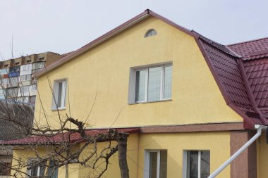 Kırmızı kiremitli çatının altında beyaz pencereli büyük sarı özel bir evin tavan arası, gri gökyüzünün arka planına karşı.