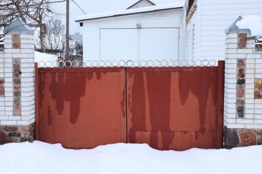 Büyük, kapalı, kırmızı, ıslak bir kapı. Tuğla duvar çitlerinin üzerinde. Kırsal kesimde kar yağışı var.