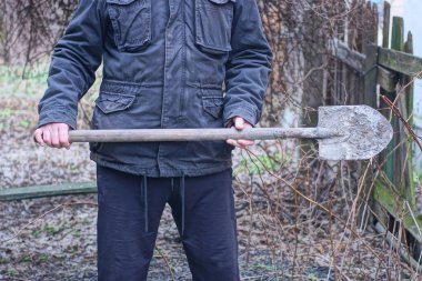 Siyah elbiseli bir erkek işçi, bir sonbahar sokağında elinde uzun, kirli bir kürek tutuyor.