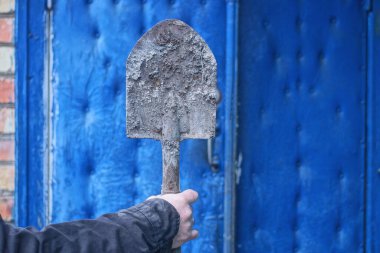 Bir elinde siyah elbiseler, gri, eski, kirli demir küreği sonbahar sokağında mavi duvara dayamış.