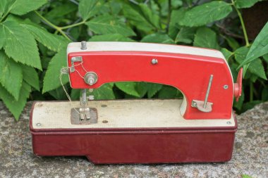 Eski kırmızı oyuncak dikiş makinesi, yaz caddesinde yeşil bitki örtüsünün yanında gri beton bir masanın üzerinde duruyor.