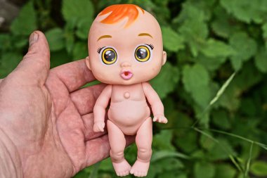 El, yeşil bitki örtüsünün arka planına karşı sokakta küçük kahverengi bir oyuncak bebek tutuyor.