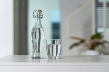 Bir bardak temiz içme suyu ve su şişesi evdeki beyaz ahşap masada..