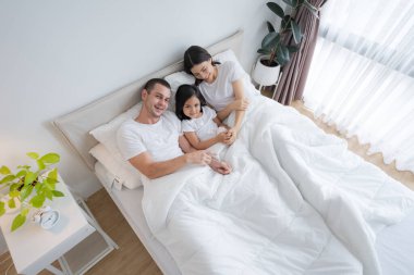 Güzel ebeveynler ve kızları mutlu gülümseyen yüzler rahat beyaz bir yatakta uzanıyorlar..