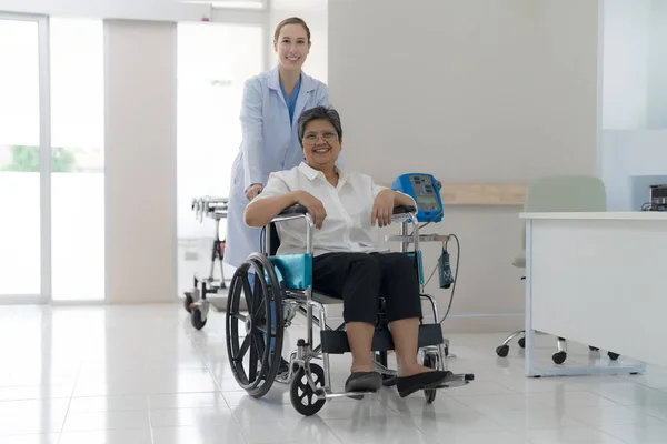 Doctor helping senior patient push wheelchair in hospital interior.