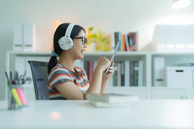 Kütüphanede gözlük ve kulaklık takan genç bir kız ya da öğrenci tablet bilgisayar kullanıyor. Gülümsüyor ve mutlu görünüyor..