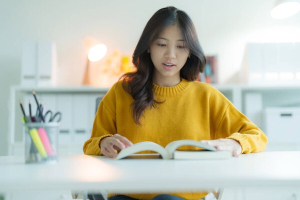 A young woman or student reading a book in the library while wearing glasses, She is smiling and looking happy.