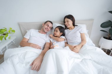 Güzel ebeveynler ve kızları mutlu gülümseyen yüzler rahat beyaz bir yatakta uzanıyorlar..