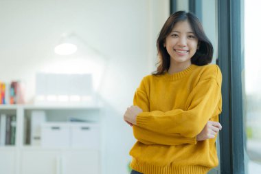 Gülümseme ve mutluluk dolu bir bakışla çekici genç bir kadın, ofisinin penceresinin önünde sıradan bir poz vererek dikiliyor..