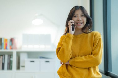 Akıllı telefon kullanan çekici genç bir kadın sıradan bir pozla pencerenin yanında durur. Ofisinde derin düşüncelere dalmış.