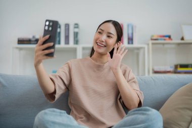 Neşeli genç bir kadın evdeki bir koltukta rahat bir şekilde oturuyor, elinde parlak bir gülümsemeyle bir cep telefonu, rahat oturma odasında kitap raflarıyla çevrili.