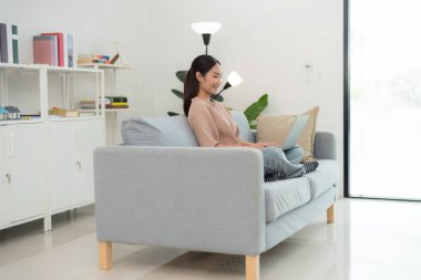 Neşeli genç bir kadın koltukta bağdaş kurarak oturuyor. Parlak, modern bir oturma odasında dizüstü bilgisayarıyla meşgul. Ev ortamında üretkenlik ve rahatlık hissi yayıyor.