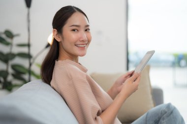 Güzel, genç Asyalı bir kadın rahat bir şekilde koltukta oturuyor, kameraya gülümsüyor ve iyi aydınlatılmış, modern oturma odasında dijital tablet kullanıyor.