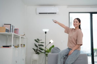 Kanepede rahat bir şekilde oturan genç kadın minimalist dekorlu, modern oturma odasındaki klimayı ayarlamak için uzaktan kumanda kullanıyor.