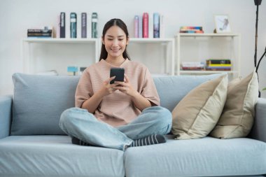 Neşeli genç bir kadın evdeki bir koltukta rahat bir şekilde oturuyor, elinde parlak bir gülümsemeyle bir cep telefonu, rahat oturma odasında kitap raflarıyla çevrili.