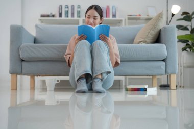 Genç kadın rahat bir kanepede, modern oturma odasında bir kitapla rahatlıyor. Minimalist bir evde huzur ve rahatlığı simgeliyor.