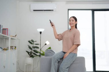 Kanepede rahat bir şekilde oturan genç kadın minimalist dekorlu, modern oturma odasındaki klimayı ayarlamak için uzaktan kumanda kullanıyor.