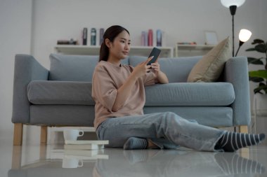 Neşeli genç bir kadın rahat bir şekilde evinde yerde oturuyor, akıllı telefonuna sıcak bir gülümsemeyle göz atıyor, gündelik bağlılık ve modern eğlence yaşam tarzını parlak bir oturma odasında şekillendiriyor.