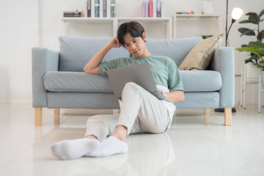 Genç Asyalı bir adam dizüstü bilgisayarına odaklanmış bir şekilde yerde oturuyor. Parlak, modern bir oturma odasında. Günlük yaşamda uzaktan çalışmayı, rahatlığı ve teknolojiyi simgeliyor.