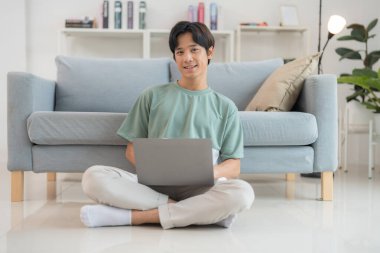 Modern oturma odasında dizüstü bilgisayarla rahat bir şekilde yerde oturan sıradan bir genç adam üretkenlik ve rahat bir yaşam tarzı sergiliyor.